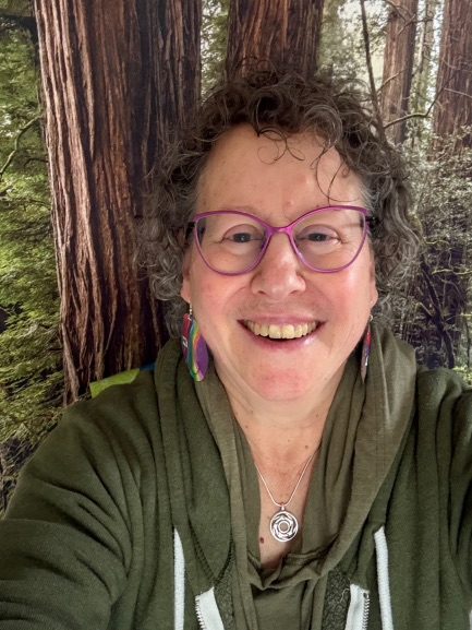 femme elder with salt and pepper curly shory hair. Smiling with purple framed glasses. Wearing a green hooded sweatshirt and a silver pendant necklace.